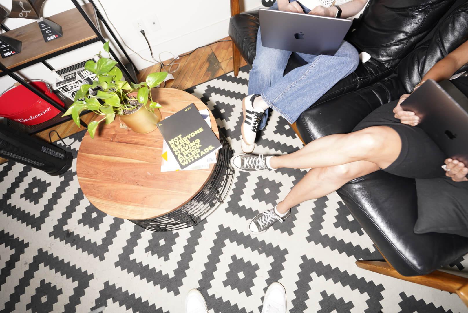 Deux personnes assises sur un canapé noir utilisant des ordinateurs portables, avec une petite table ronde tenant une plante et un livre, sur un tapis à motifs.