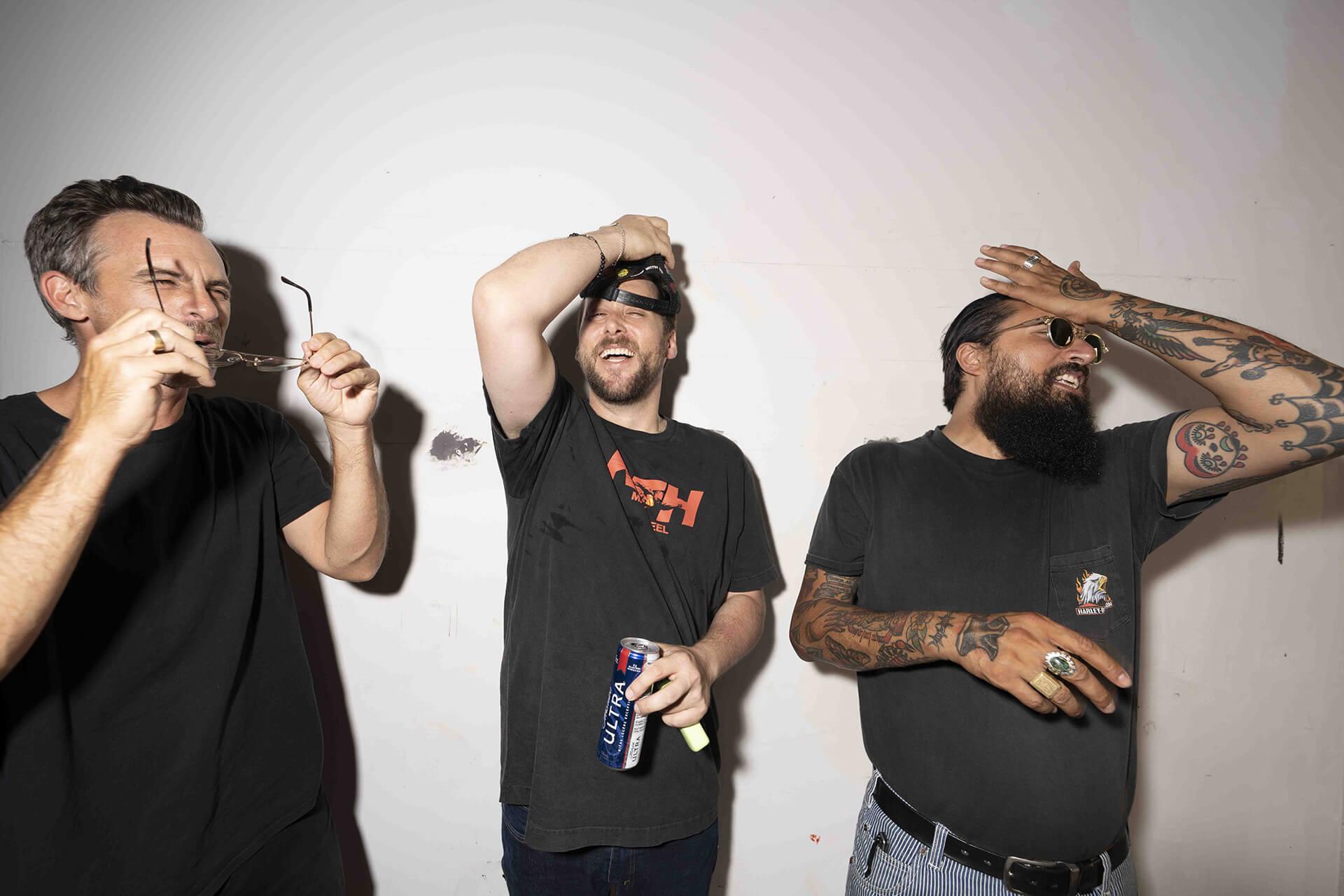 Trois hommes se tiennent contre un mur, riant et se couvrant le visage.