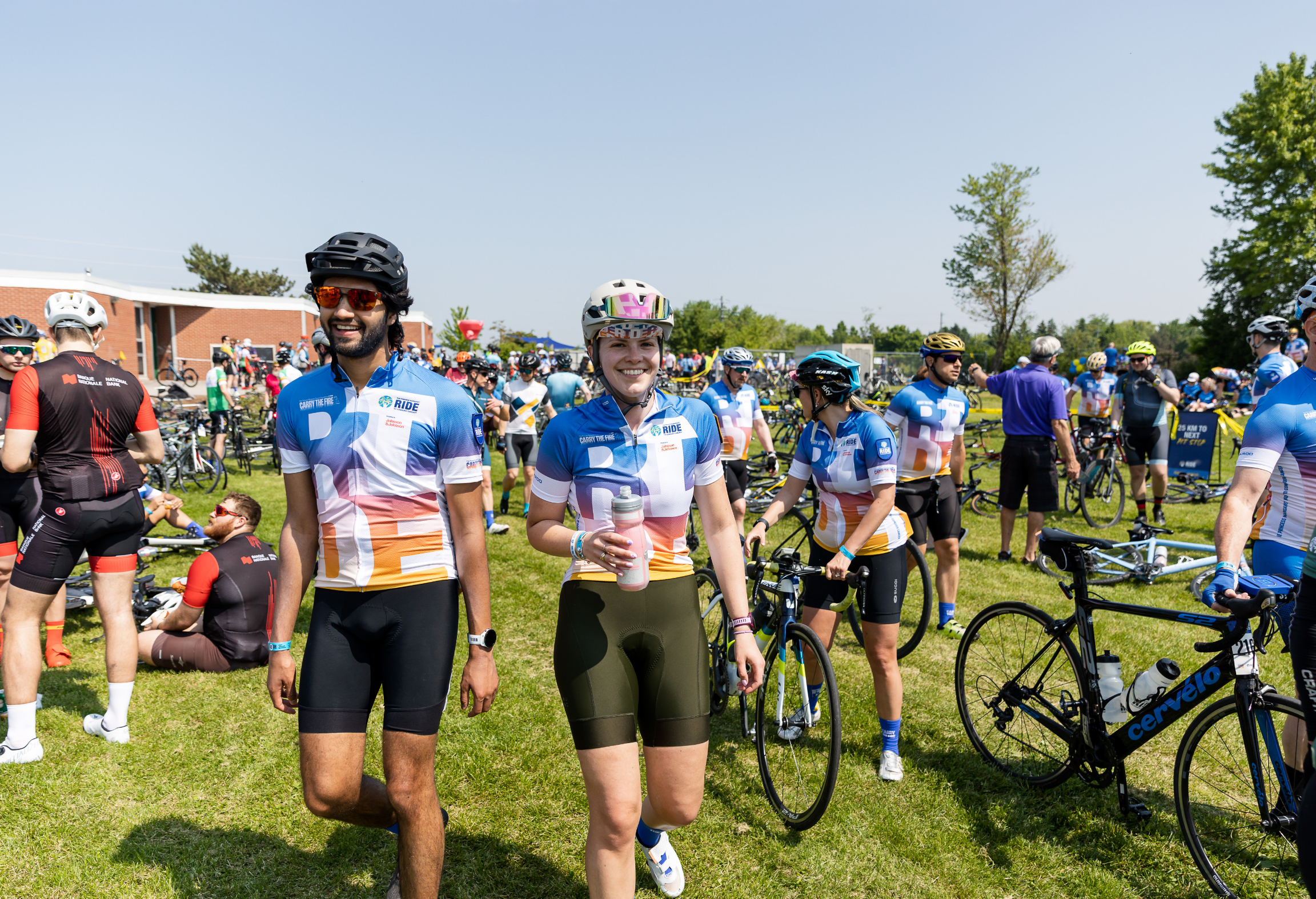 Des cyclistes en maillots colorés se rassemblent sur un terrain herbeux, certains tenant des vélos et d'autres debout ou assis.