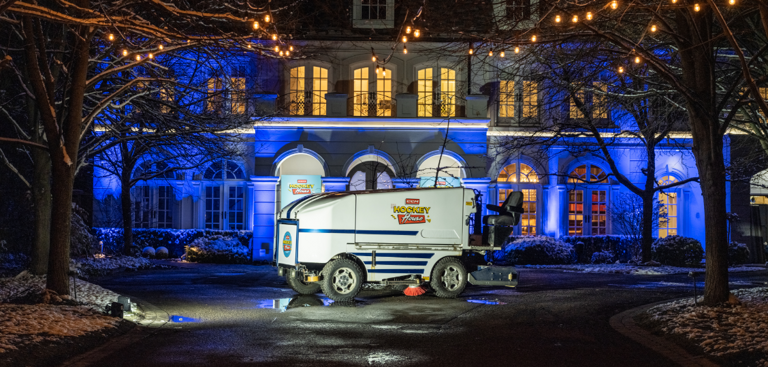 A lit-up ice resurfacing machine parked in front of a large, illuminated building at night.