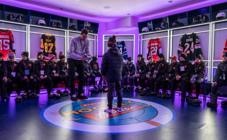 A group of children in winter clothing sit in a brightly lit locker room with hockey jerseys on display.