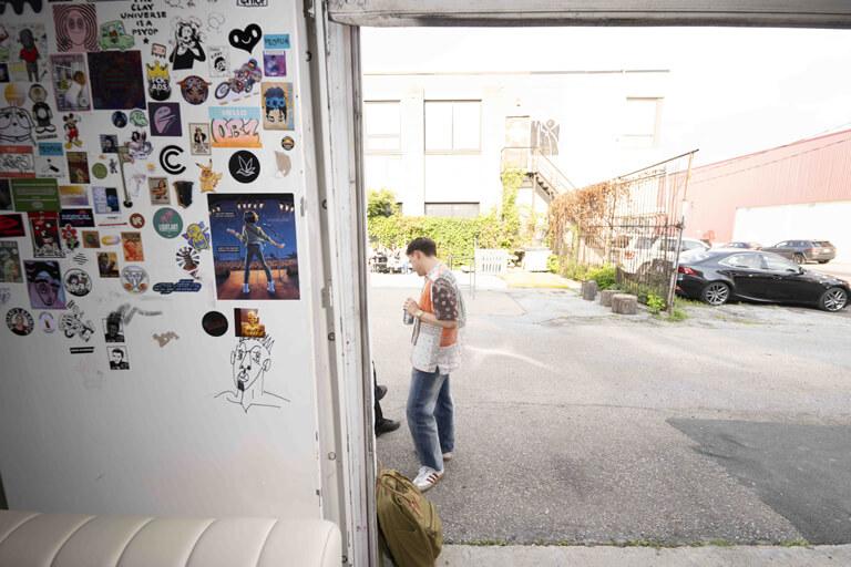 Une personne se tient dehors dans une zone de stationnement, vue à travers une porte ouverte avec un mur couvert de divers autocollants et affiches.