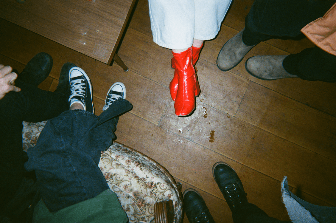 Un groupe de personnes debout en cercle avec diverses chaussures, y compris des bottes rouges vives, sur un plancher de bois.