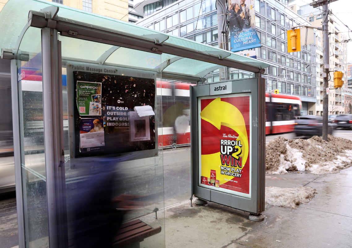 A city bus stop with advertisements and a trolley passing by, set against a backdrop of urban buildings.