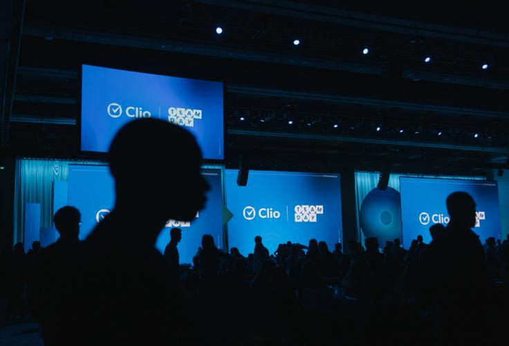Silhouettes of people in front of large blue screens displaying "Clio Team Day" at an event.