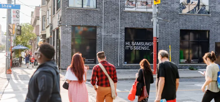 People crossing a street in front of a brick building with posters and advertisements.