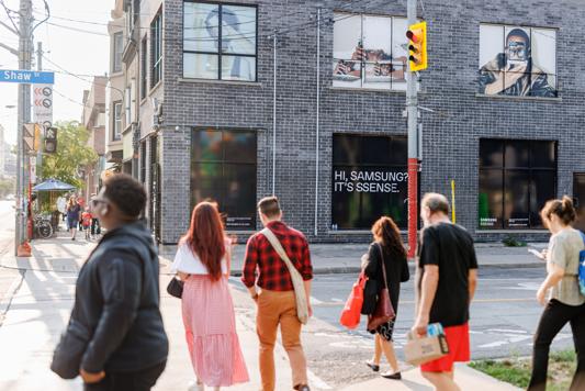 People crossing a street in front of a brick building with posters and advertisements.