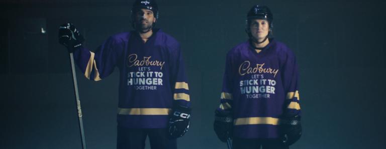 Deux joueurs de hockey portant des maillots violets avec « Cadbury, Let's Stick It To Hunger Together » imprimé dessus.