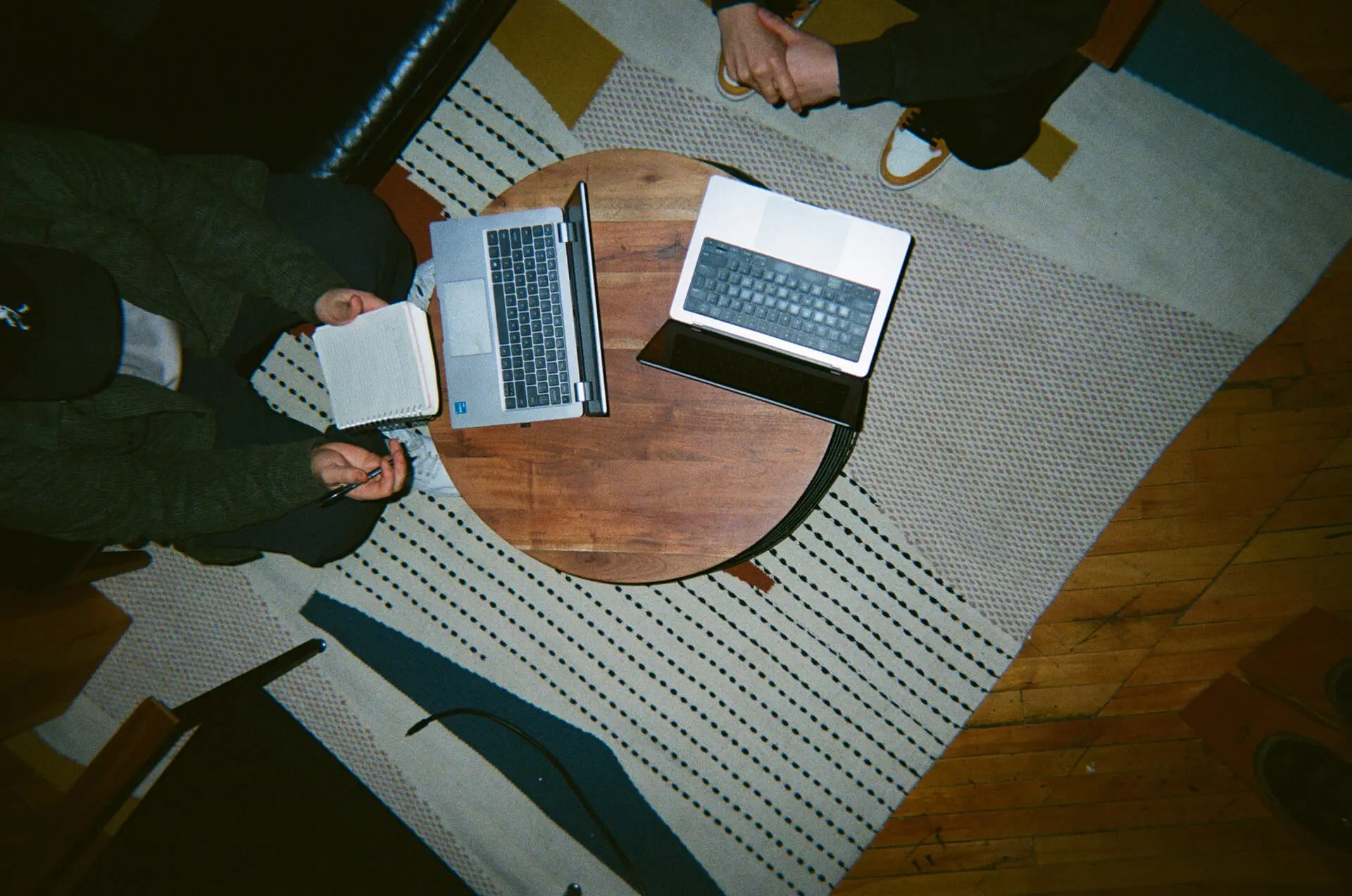 Overhead view of two people sitting at a round table with laptops and a notebook on a patterned rug.