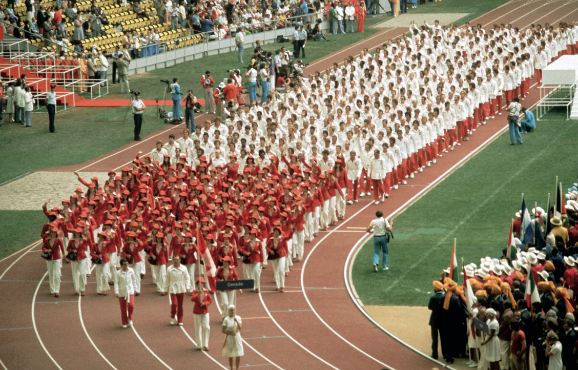 A large group of athletes in red and white uniforms march together in a stadium during an opening ceremony.