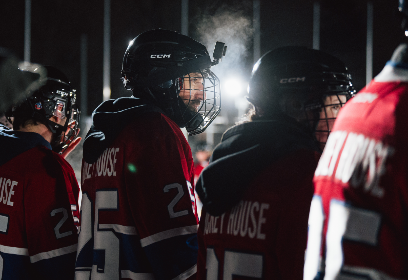 Hockey players wearing red jerseys and helmets stand on the ice, with visible breath in the cold air.