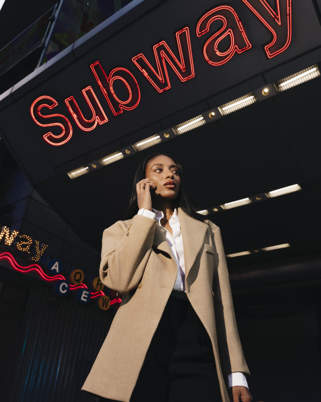 A woman in a tan coat is talking on the phone under a subway sign.