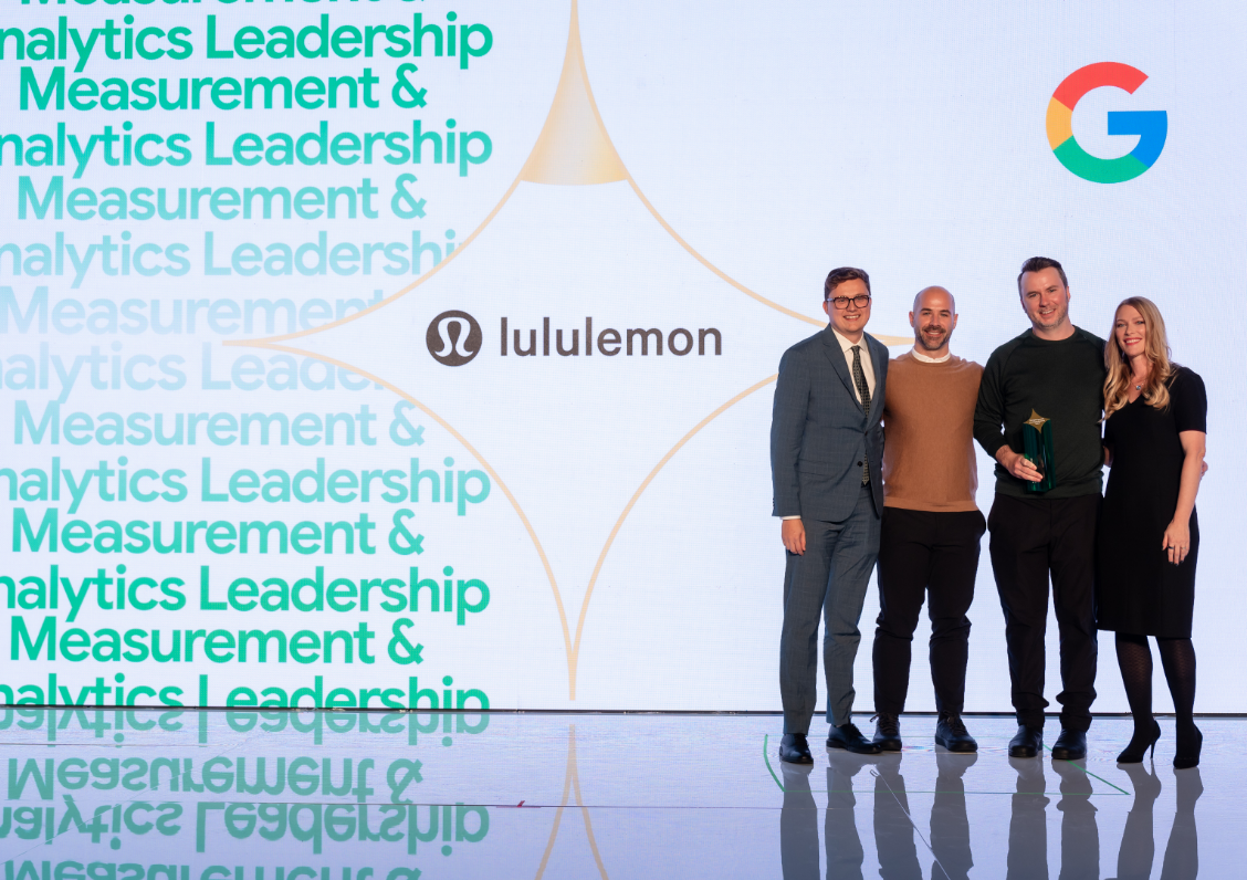 Four people pose on stage with a Lululemon logo and Google symbol on a large screen behind them.