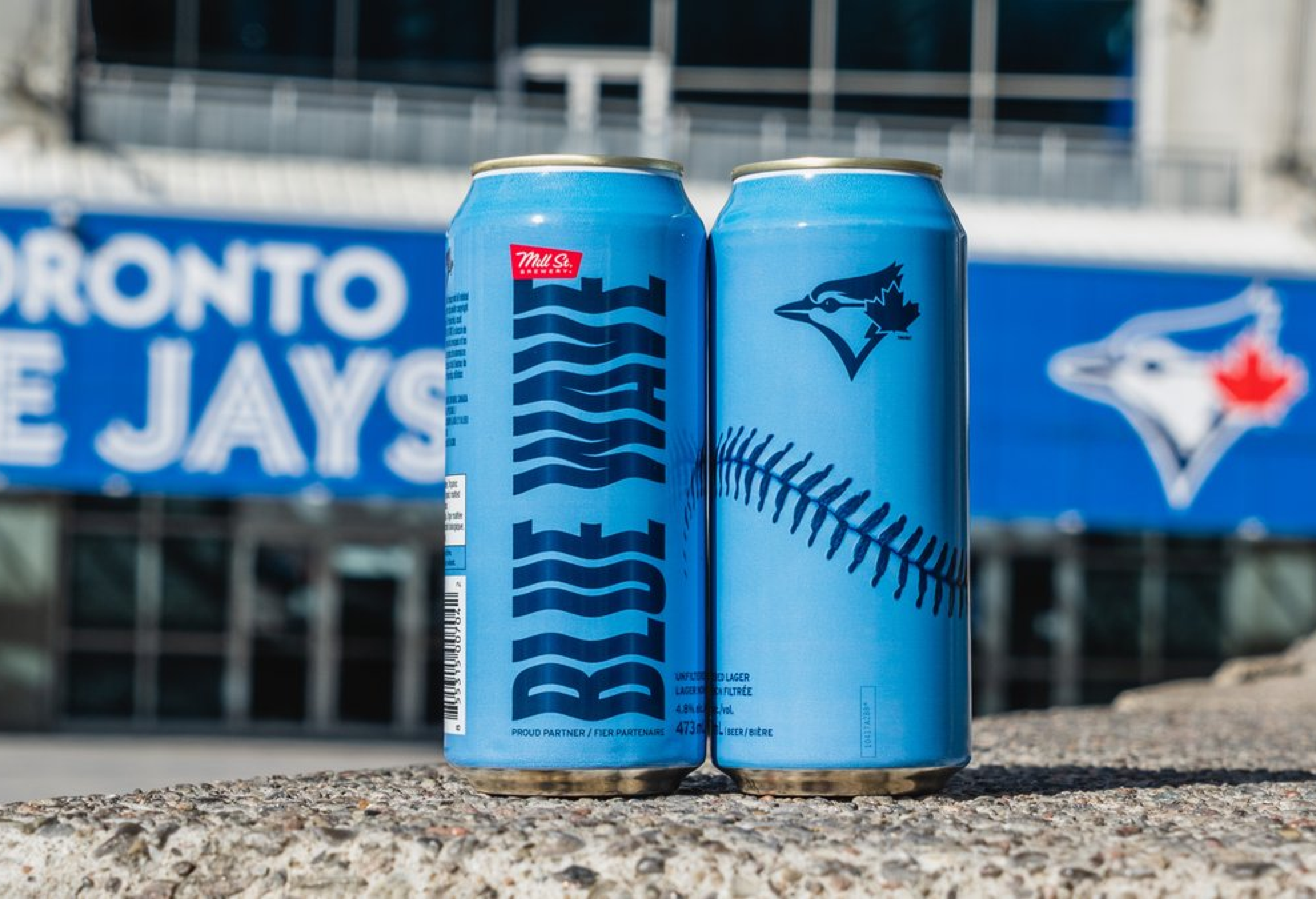Two blue cans with a baseball-themed design in front of a Toronto Blue Jays sign.