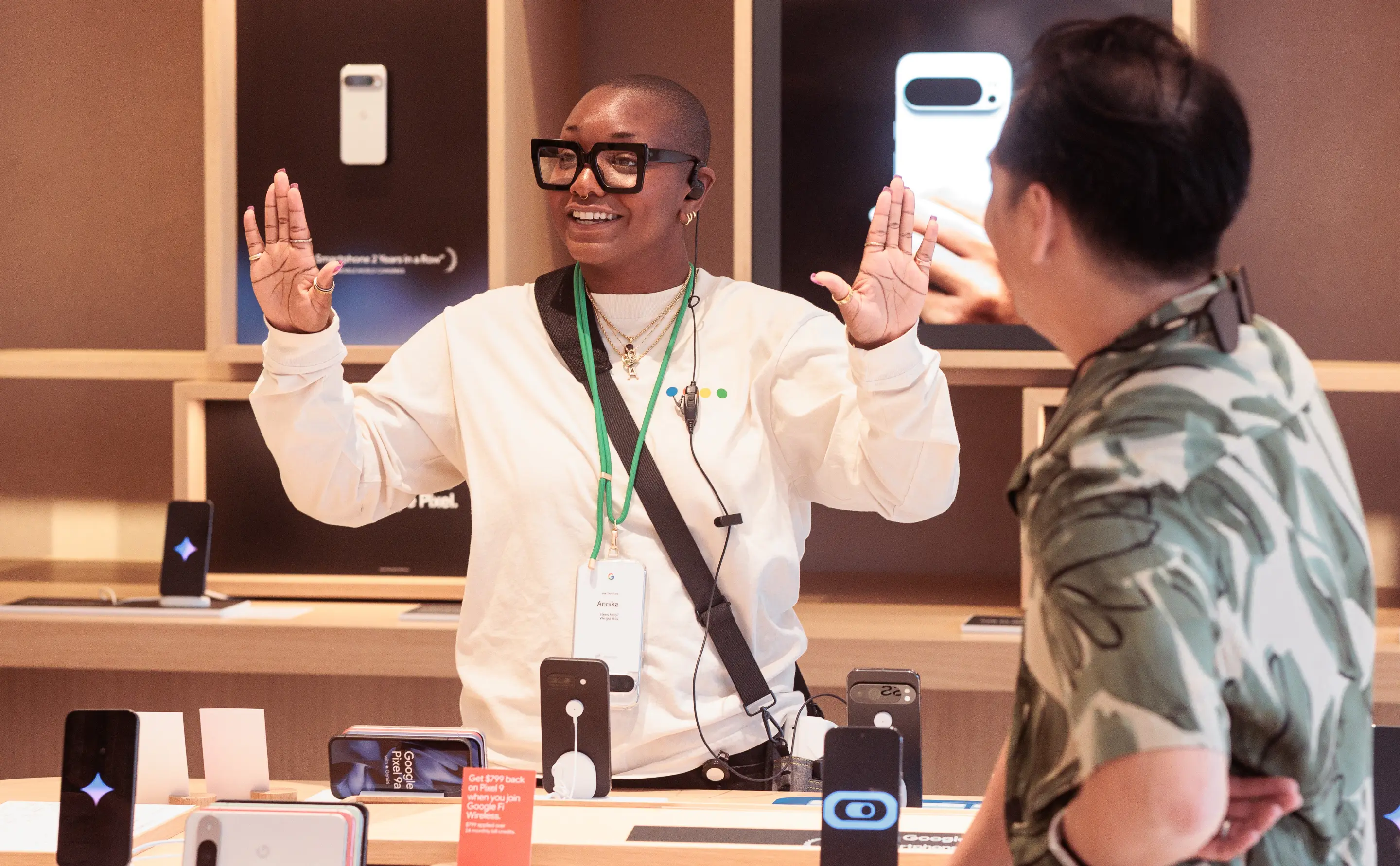 Une personne portant des lunettes et un badge parle avec enthousiasme à une autre personne dans un magasin de technologie.