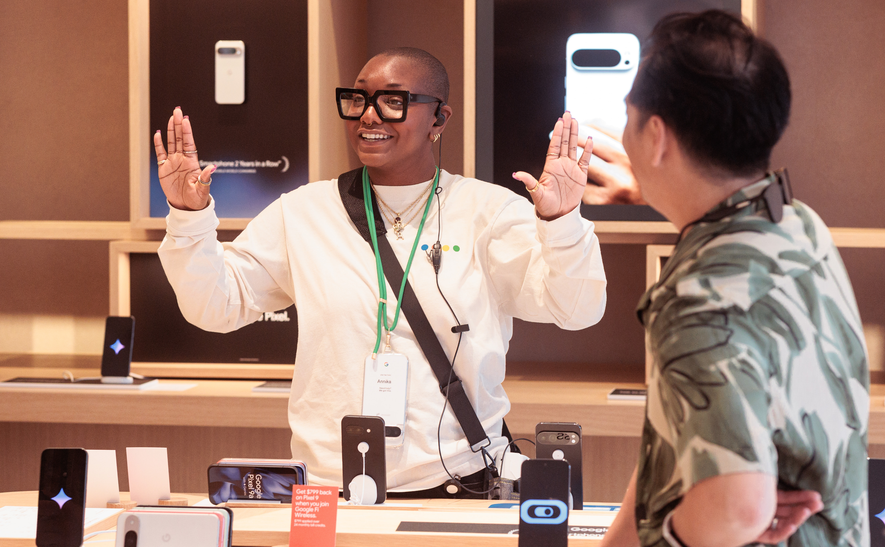 A person wearing glasses and a nametag is enthusiastically speaking to another person in a tech store.