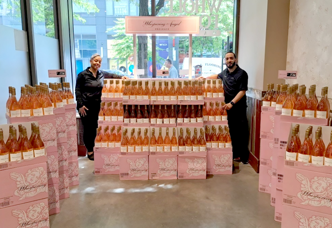Two people standing beside a large display of Whispering Angel wine bottles in a store.