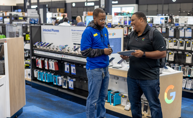 Two men are talking and looking at a tablet in an electronics store.