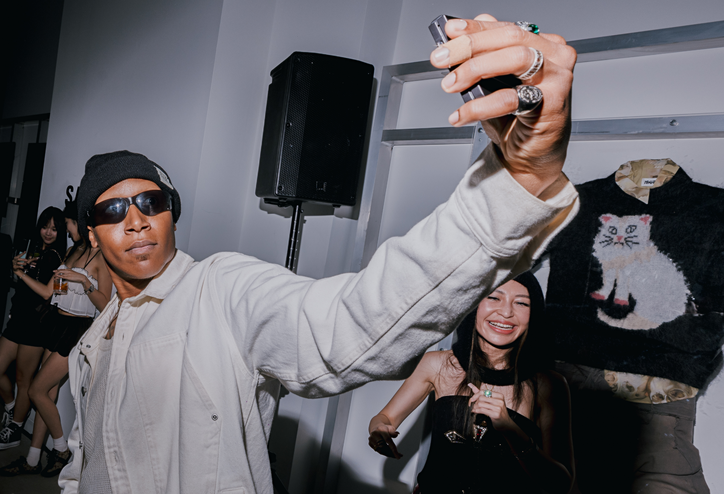Two people taking a selfie at a party, with a sweater featuring a cat design in the background.
