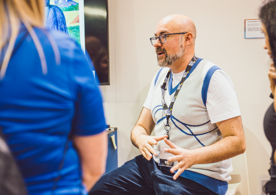 A man wearing glasses and a lanyard is speaking to a small group of people.