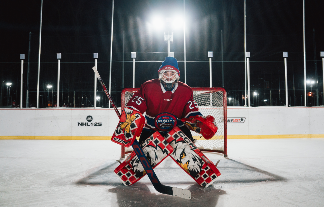 A hockey goalie in full gear stands in front of the net on an outdoor ice rink at night.