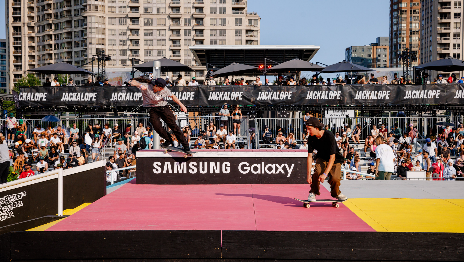 Deux skateurs effectuant des figures sur une rampe lors d'un événement en plein air avec une foule qui regarde.