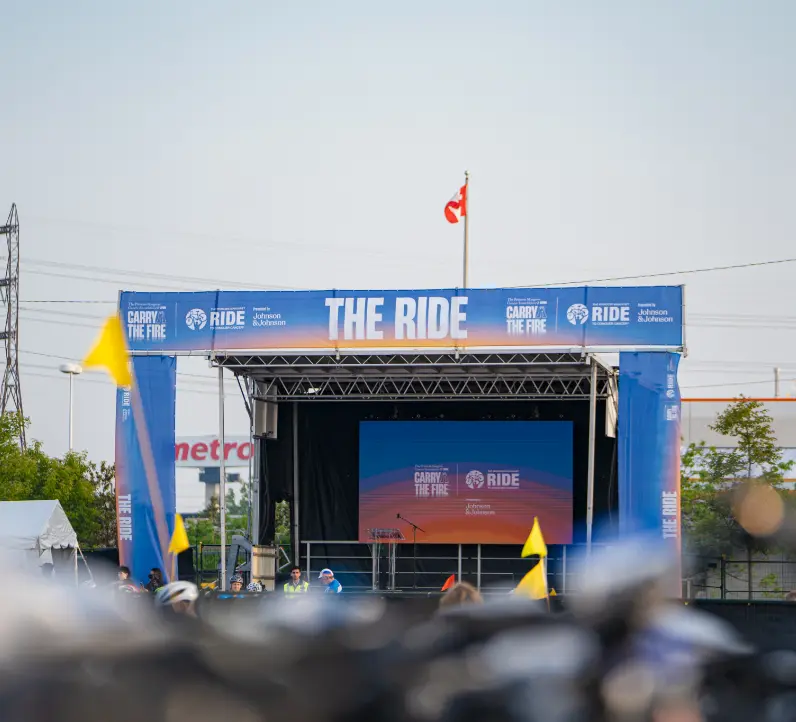 Scène extérieure avec un grand écran et une bannière « The Ride », entourée de quelques personnes et de drapeaux jaunes.