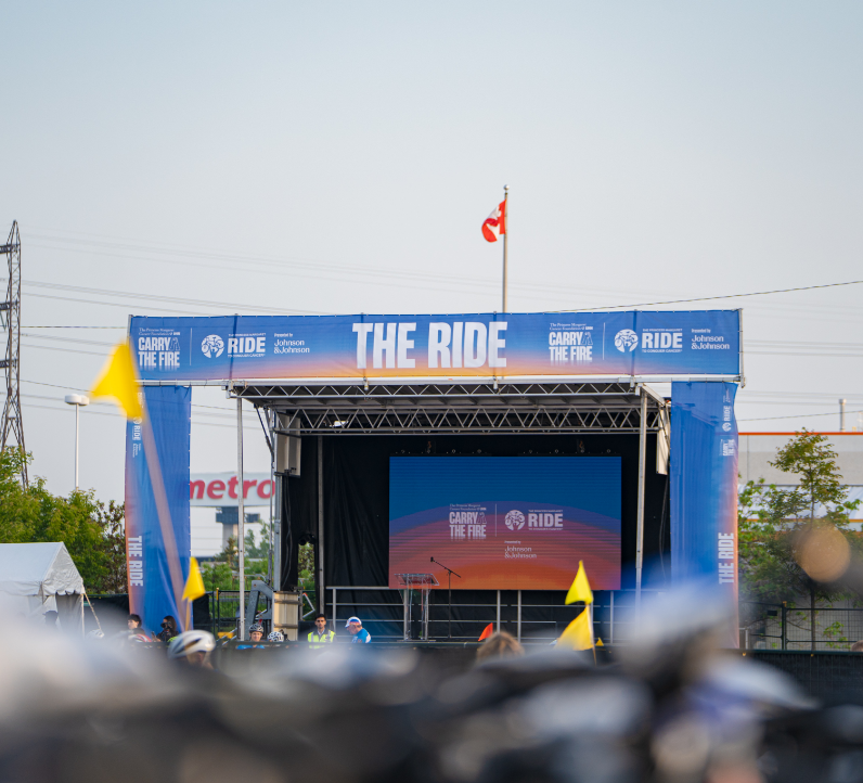 Scène extérieure avec un grand écran et une bannière « The Ride », entourée de quelques personnes et de drapeaux jaunes.
