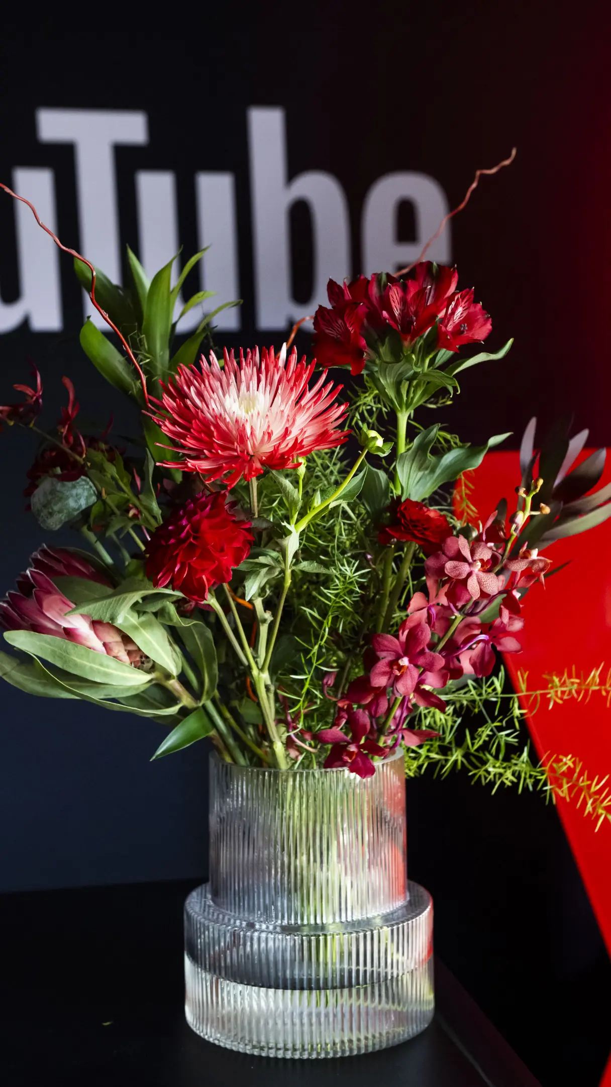 A vibrant bouquet of red and pink flowers in a clear ribbed vase, with a partially visible logo in the background.