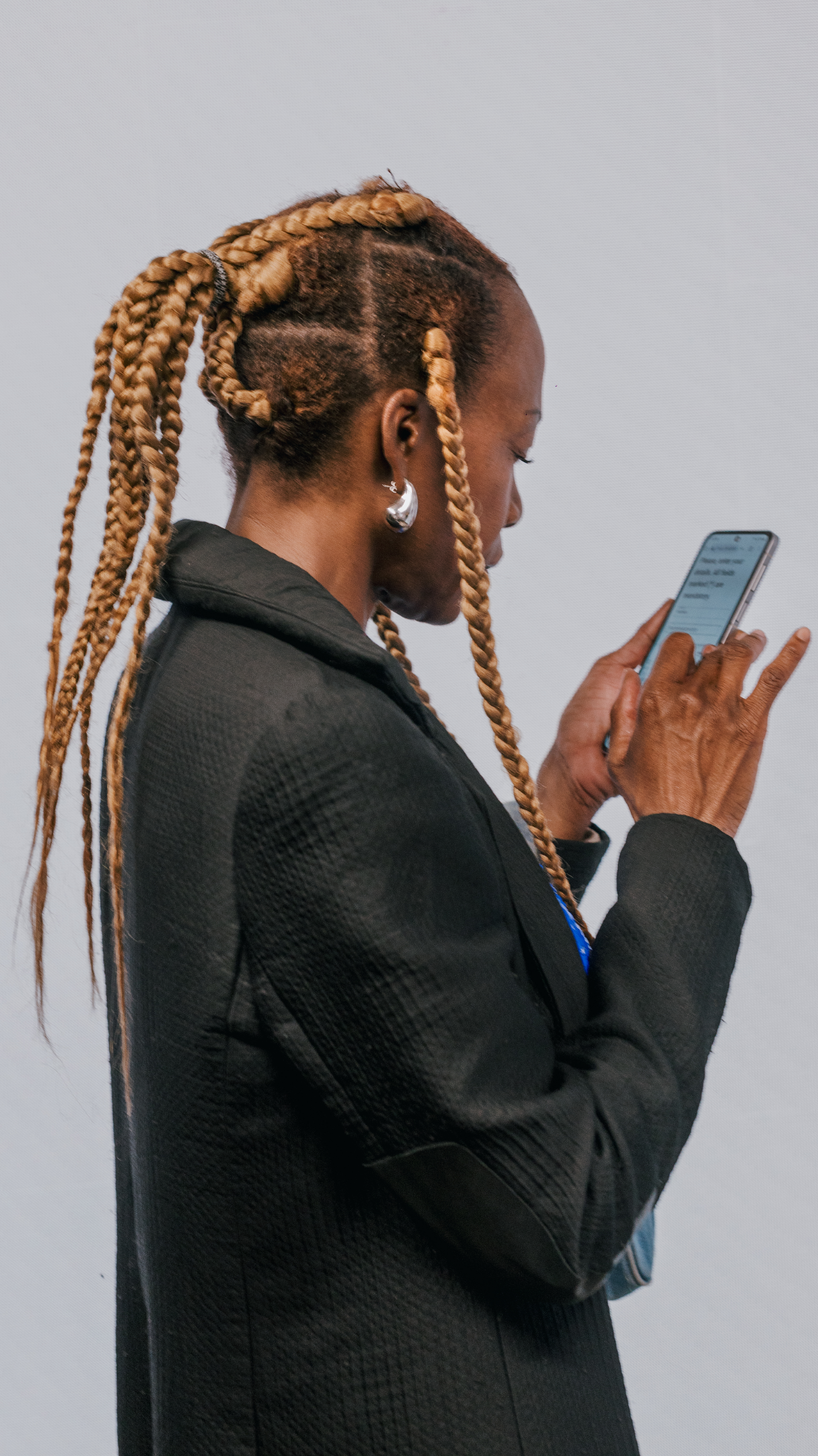 A person with braided hair is wearing a black jacket and looking at a phone.