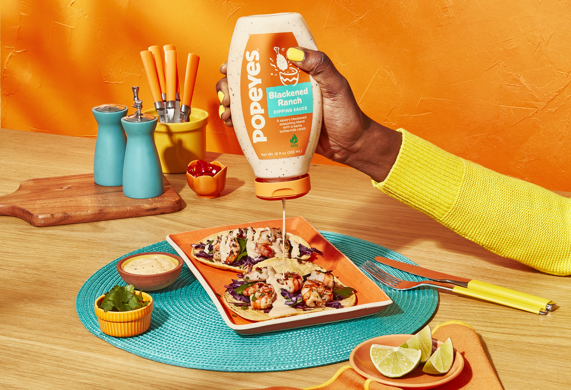 A hand is pouring Popeyes Blackened Ranch sauce onto tacos on a plate on a colorful table setting.