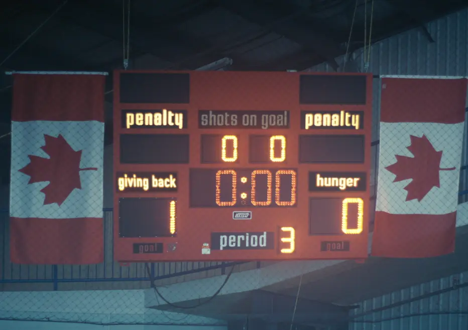 Un tableau de pointage de hockey montre « Giving Back » menant « Hunger » 1-0 avec 0:00 restant en troisième période, flanqué de drapeaux canadiens.