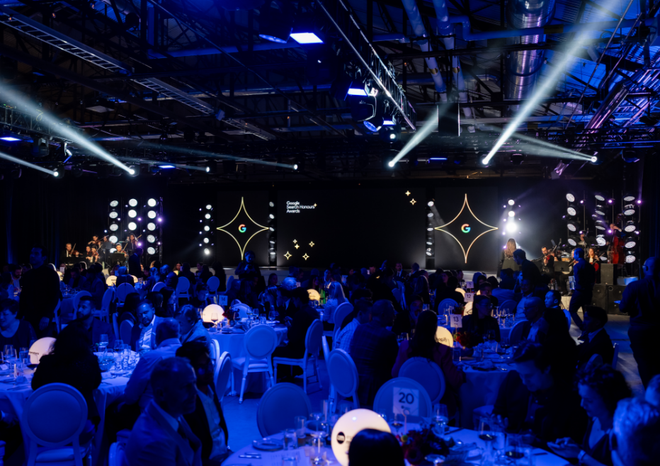 A large event with people seated at tables, illuminated by overhead and stage lighting.