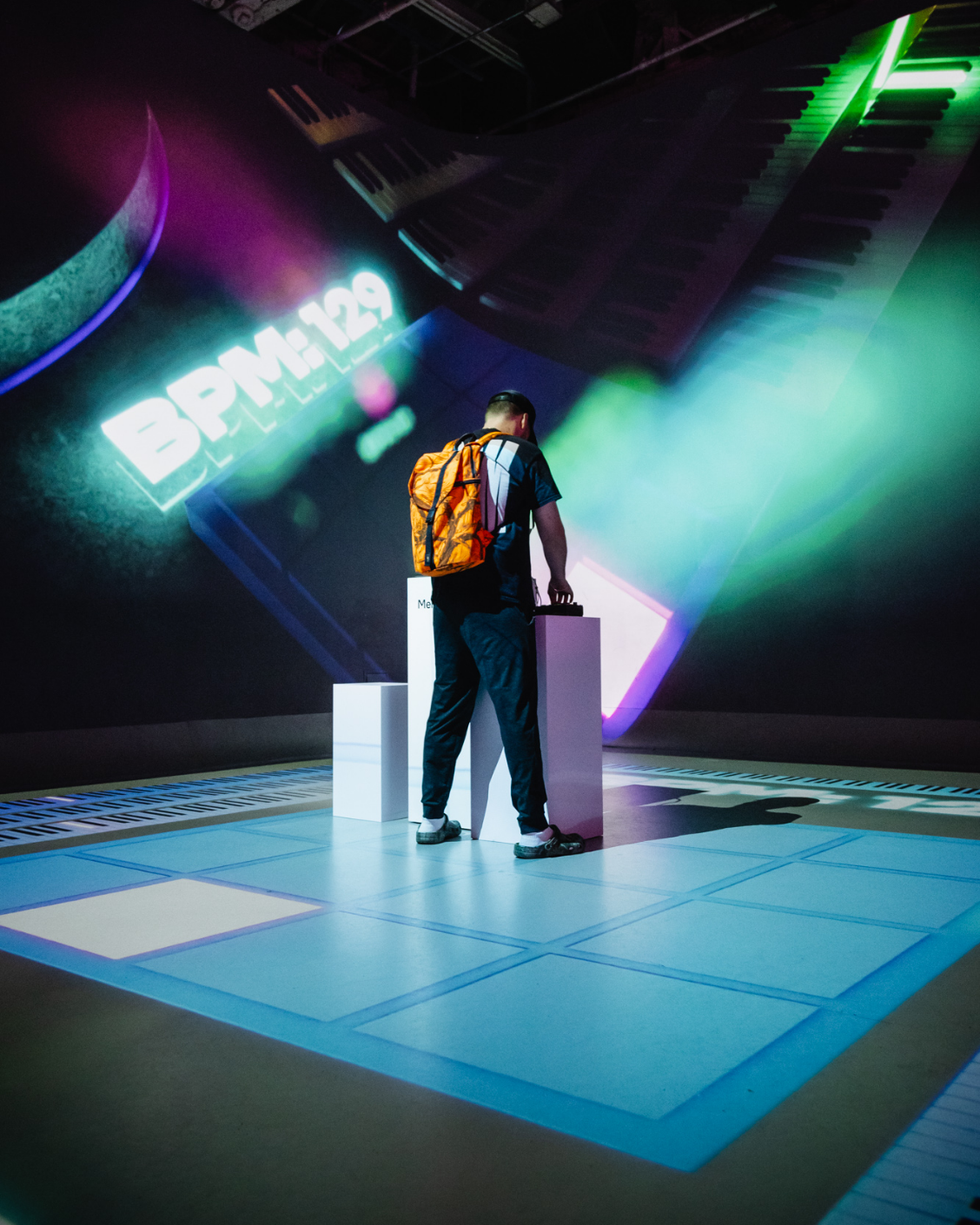 A person with an orange backpack interacts with an exhibit under colorful lights, with "BPM: 129" displayed in the background.