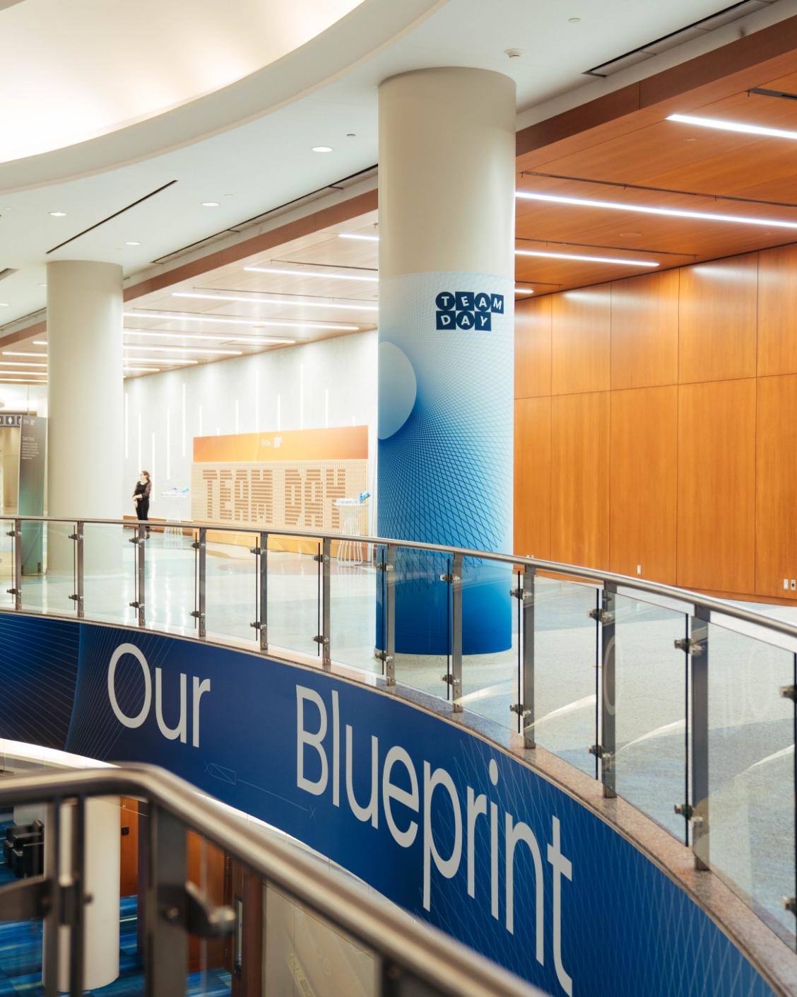 A modern, curving hallway with signs reading "Team Day" and "Our Blueprint" in a spacious building interior.