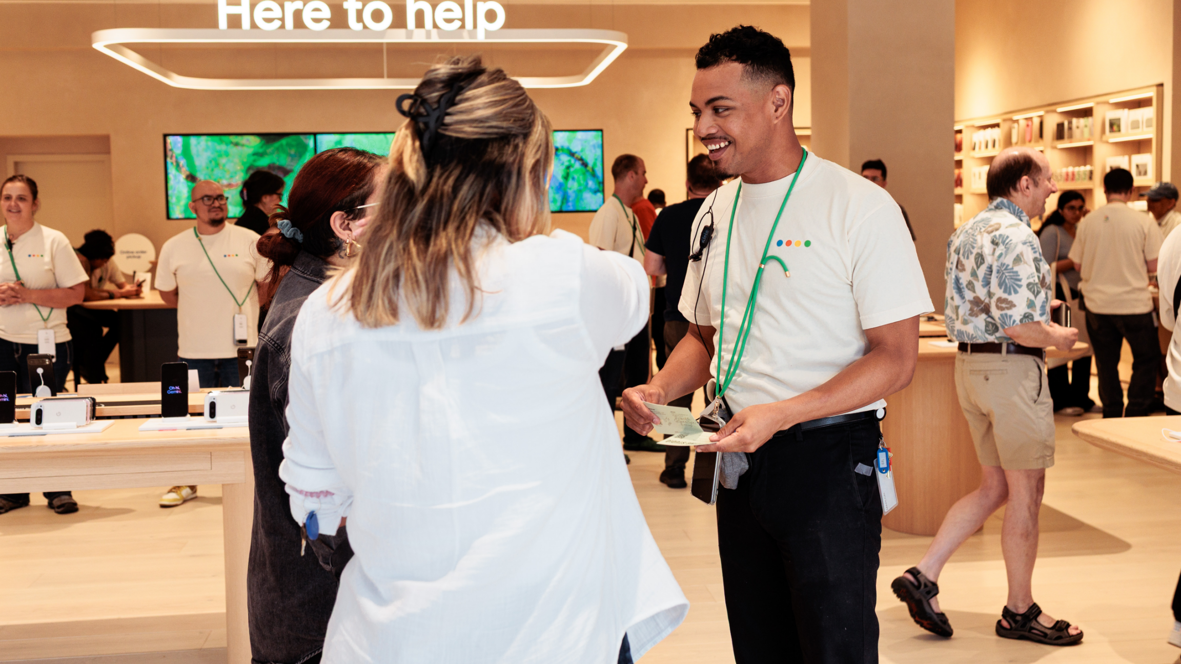 People interacting inside a busy electronics store with a "Here to help" sign above.