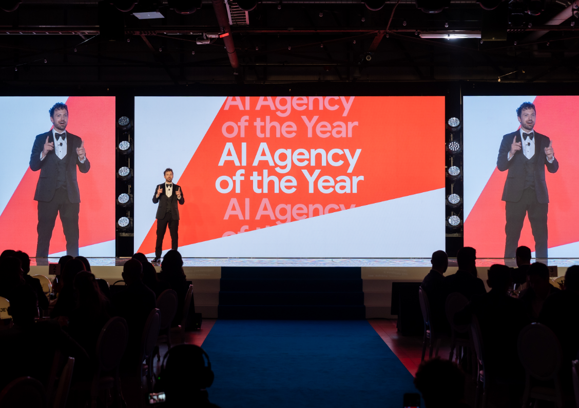 A person in formal wear speaks on stage at an "AI Agency of the Year" event, with the title displayed on large screens.
