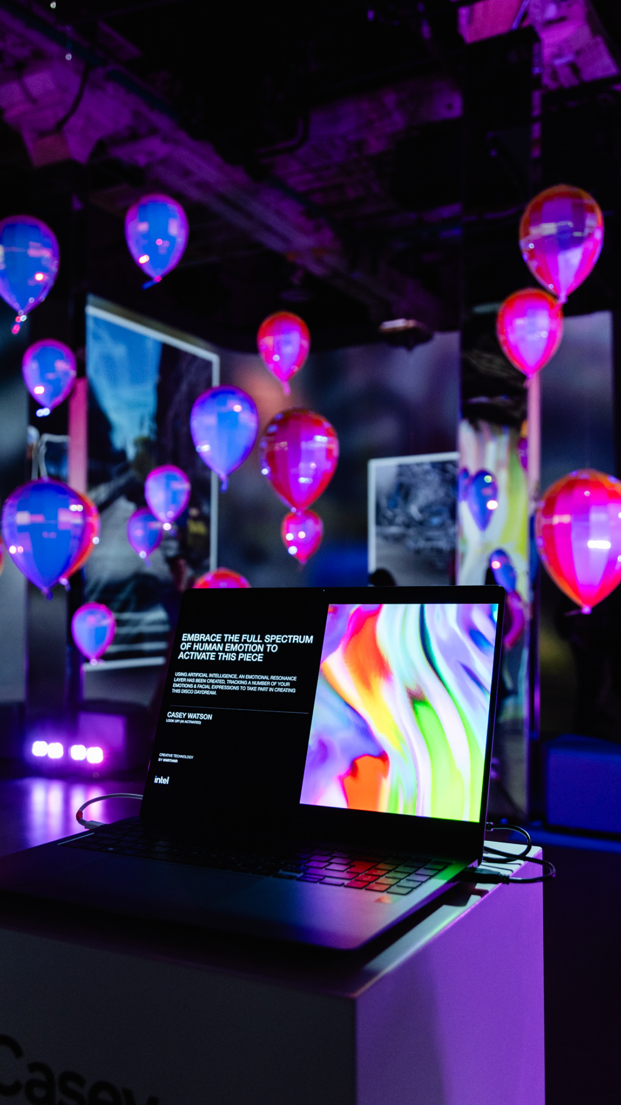 A laptop displaying colorful digital art is set in a room with glowing balloons and vibrant lights.