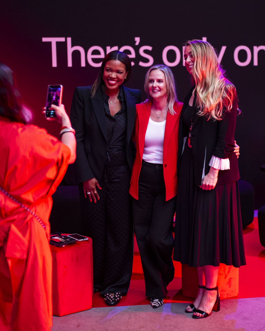 Three women pose for a photo together at an event with a "There's only on" sign in the background.