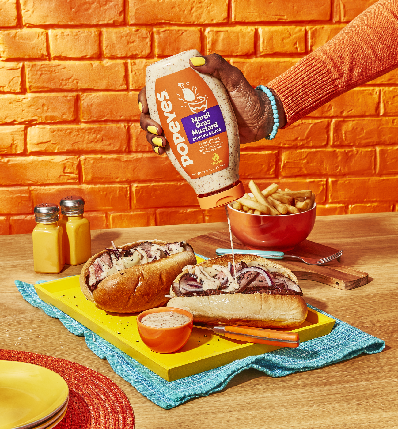 A person is pouring Mardi Gras Mustard sauce onto sandwiches on a wooden table, next to fries and condiments.