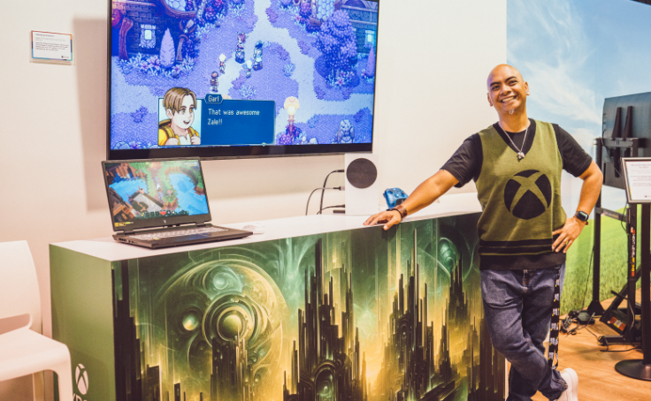 Une personne souriant et debout à côté d'une configuration de jeu avec un jeu vidéo affiché sur un moniteur et un ordinateur portable.