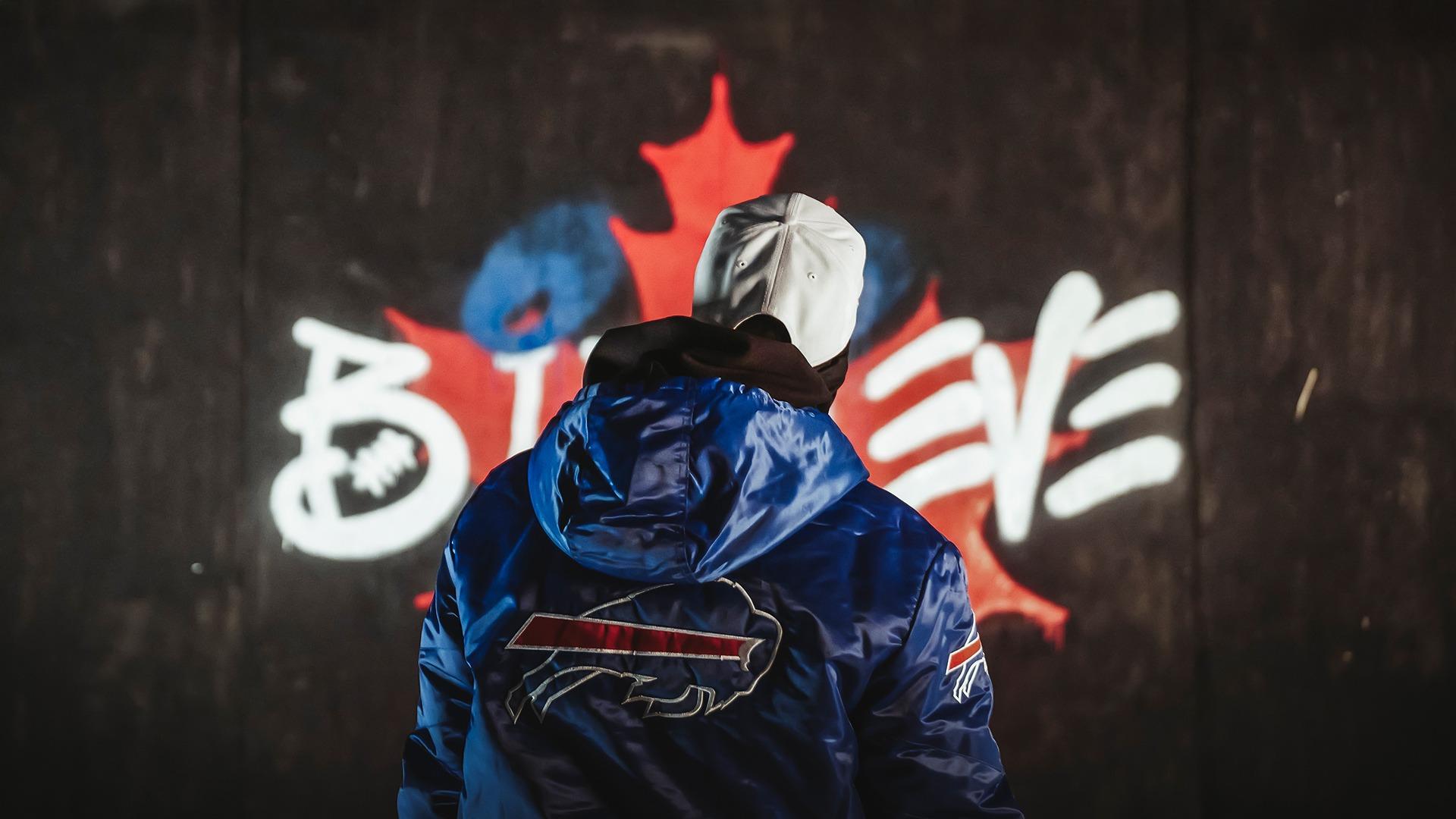 A person in a blue jacket with a buffalo emblem stands in front of graffiti featuring a maple leaf.