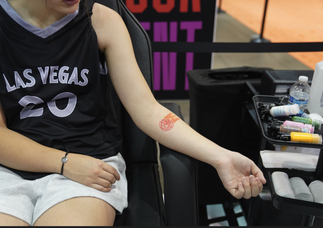 A person wearing a "Las Vegas" jersey displays a red temporary tattoo on their arm while sitting next to a tray of tattoo supplies.