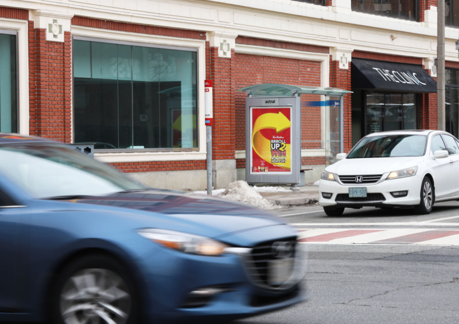 Cars passing by a bus stop in front of a brick building on a city street.