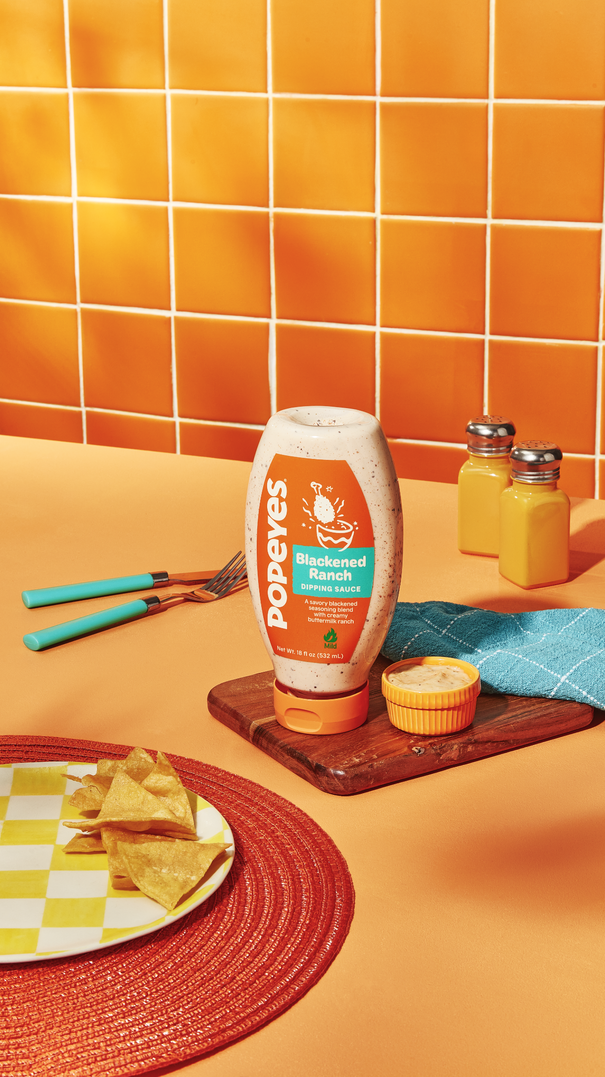 Une bouteille de sauce trempette Popeyes Blackened Ranch sur une table avec des chips, des ustensiles et des condiments sur un fond de carreaux orange.