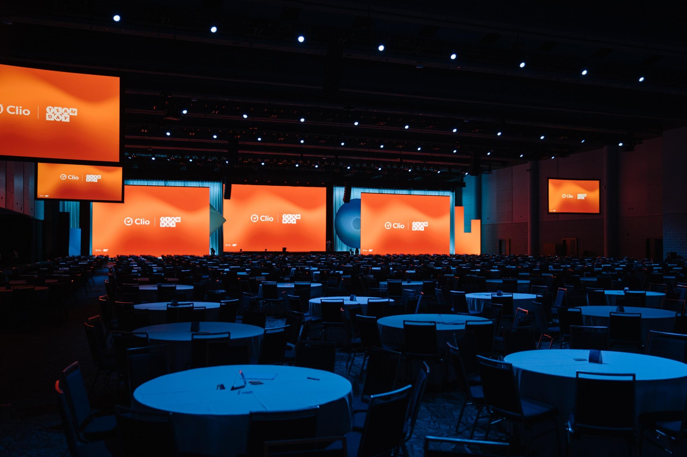 A large, dimly lit conference room with multiple round tables and bright orange screens displaying "Clio" and "Team Day."