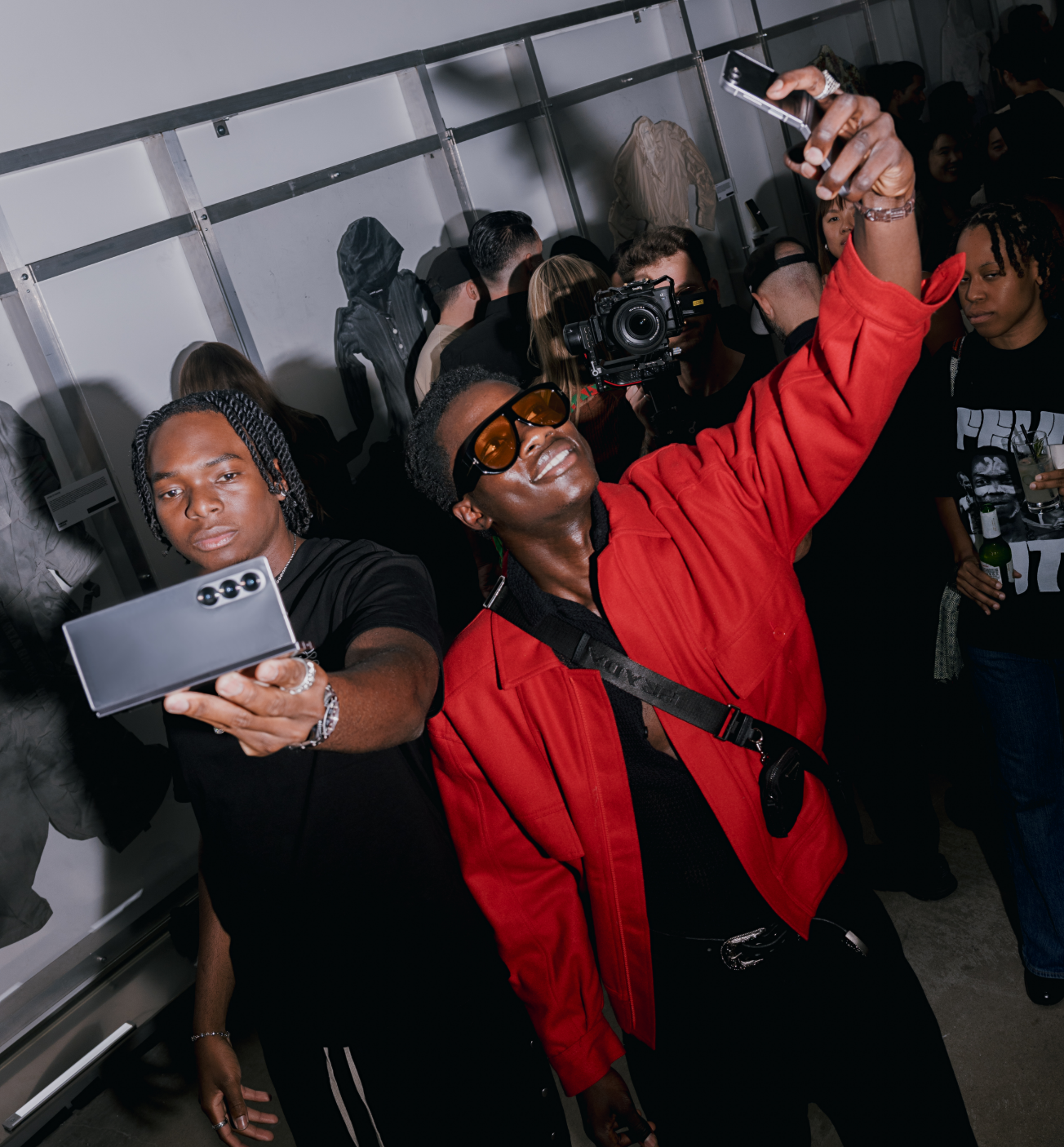 Two people are taking selfies at a crowded event, with others in the background, including someone holding a camera.