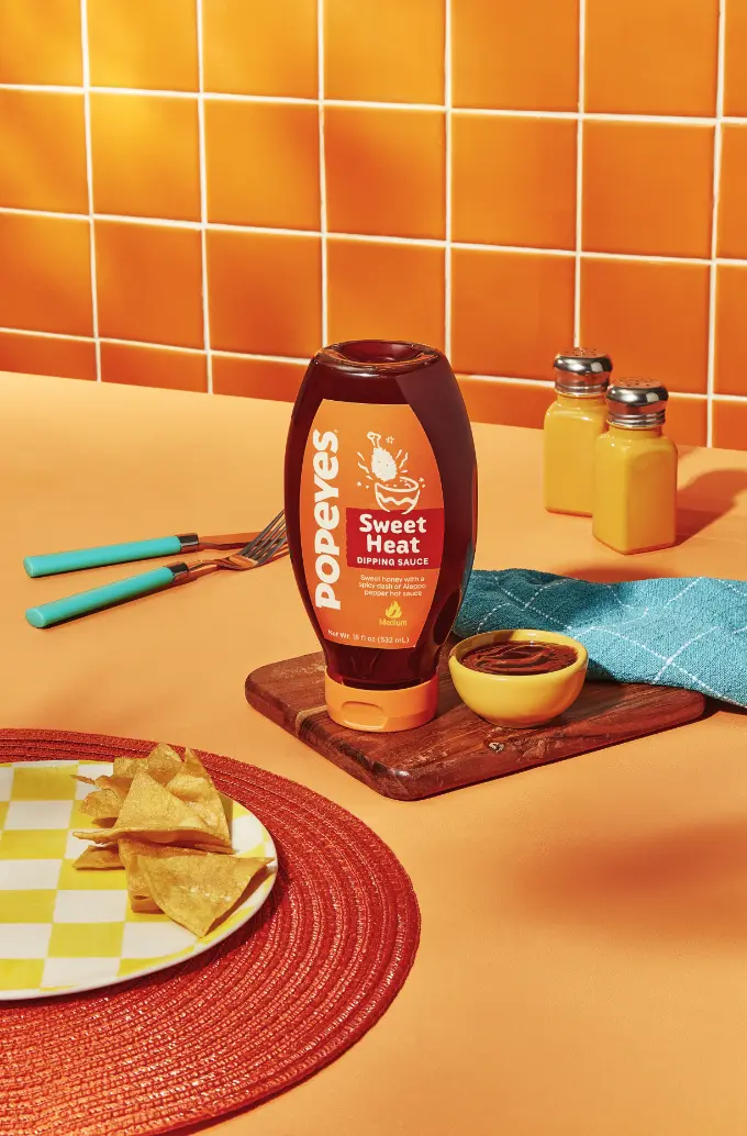 A bottle of Popeyes Sweet Heat dipping sauce is placed on a wooden board next to a small bowl of sauce, with tortilla chips on a checkered plate and an orange tiled background.