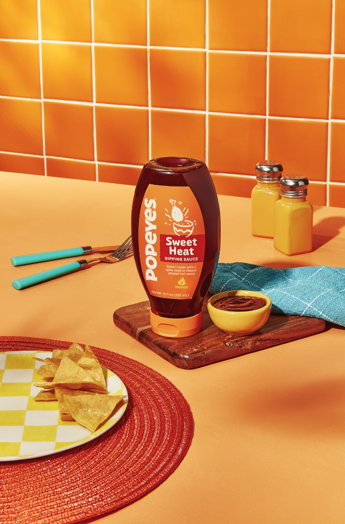 Une bouteille de sauce trempette Popeyes Sweet Heat est placée sur une planche en bois à côté d'un petit bol de sauce, avec des chips de tortilla sur une assiette à carreaux et un fond de carreaux orange.