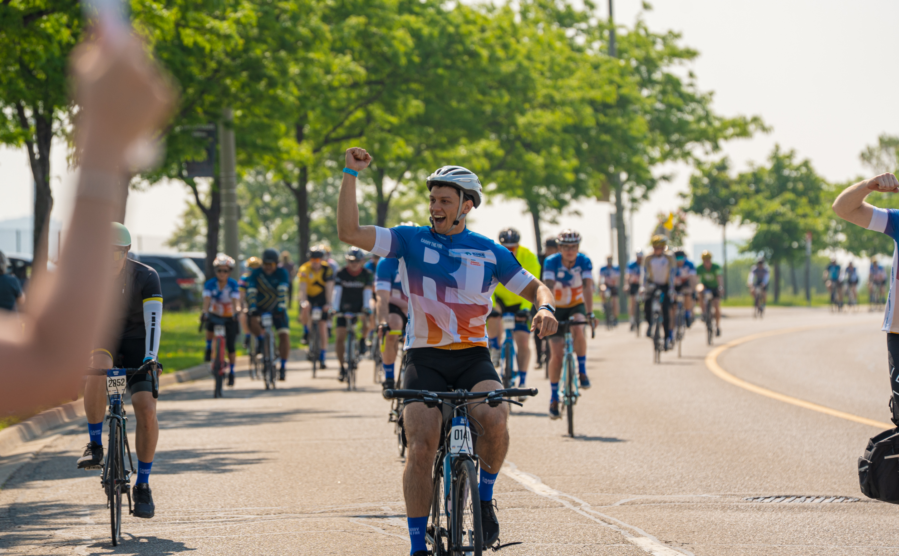 A group of cyclists rides together on a sunny road with trees lining the path, and one cyclist in the foreground raises an arm triumphantly.